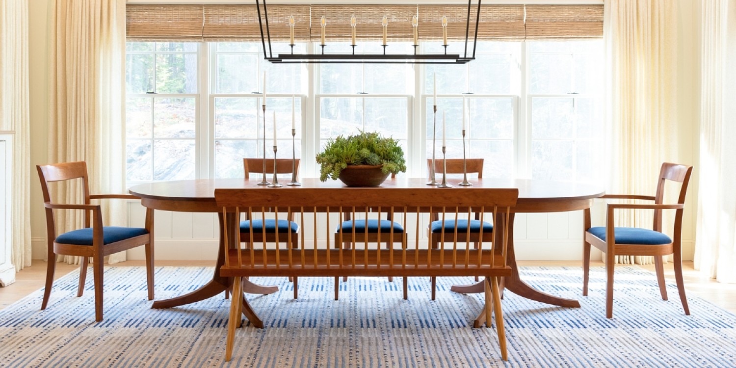 Dining room with yellow walls and a full wall of windows behind a double pedestal table. Around the table are wooden chairs and a wooden spindle bench sits in front of the table. A cast iron candle chandelier hangs from the ceiling.