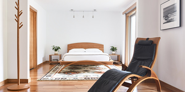 Bedroom scene. A wooden bed with a curved footboard is sitting on a light colored rug. The bed has white linens and pillows. On either side of the bed are wooden cube nightstands with potted plants. To the left is a wooden coat tree. On the right is a wooden chaise lounge chair with black leather upholstery with a black and white photograph hanging on the wall. On the floor is a woven rug.