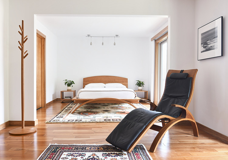 Bedroom scene. A wooden bed with a curved footboard is sitting on a light colored rug. The bed has white linens and pillows. On either side of the bed are wooden cube nightstands with potted plants. To the left is a wooden coat tree. On the right is a wooden chaise lounge chair with black leather upholstery with a black and white photograph hanging on the wall. On the floor is a woven rug.