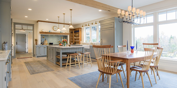 Interior of an open concept kitchen and dining room. The dining room is in the foreground where a wooden dining table and wooden spindle chairs sit on top of a blue rug. There is a blue kitchen island with wooden crescent stools and a kitchen with blue cabinetry in the background.