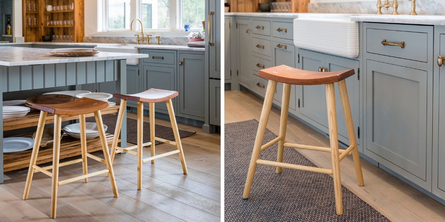 Two Images. The left is two wooden crescent stools at a kitchen counter. There is shelving with white serving dishes and on the marble counter is a wooden bowl. The image to the right is a close up of a crescent stool beside a blue kitchen cabinet with brass pulls and a deep white kitchen sink. The stool is sitting on a blue and cream woven rug and oak floorking.