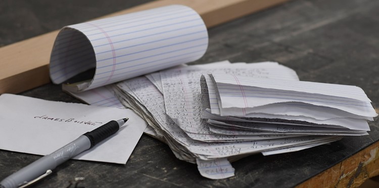 A lined pad of paper sits on a woodworkers workbench. It is flipped open with several loose sheets of paper that have dates and furniture codes written on them. To the left there is an envelope with a name on it and a pen with the cap off.