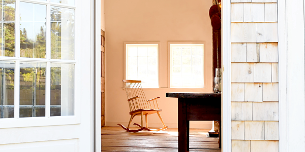 Looking inside a house from outside a doorway. There is a rocking chair in the far corner and in the foreground is a large dark wood table. The house has cedar shingles and there are a few purple chive blossoms in bloom in the lower right hand corner.