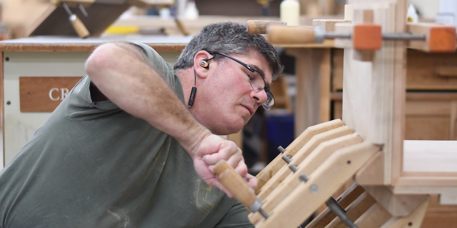 A craftsman works on a piece of furniture in a workshop. He is using wooden clamps to hold a piece of wood in place.