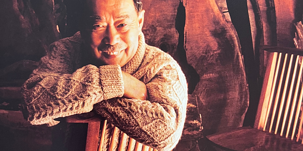 A picture of George Nakashima. He is sitting in one of his spindle chairs and wearing a cream colored woven sweater and light pants. Behind him are large live-edge slabs of wood.