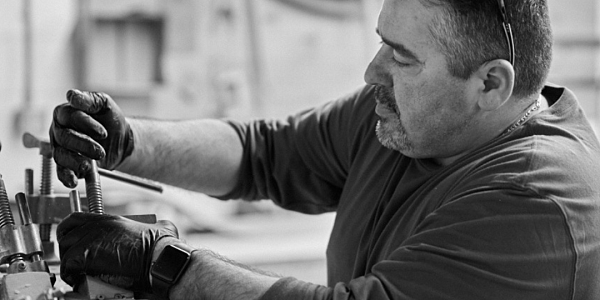 A man works on turning a clamp. He is wearing a dark shirt and has glasses on his head.