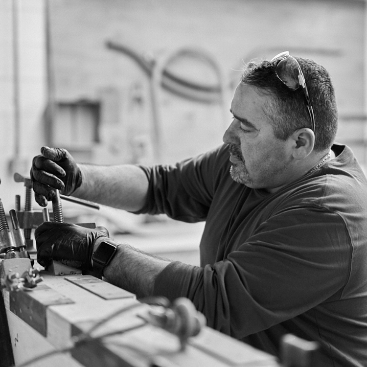 A man works on turning a clamp. He is wearing a dark shirt and has glasses on his head.